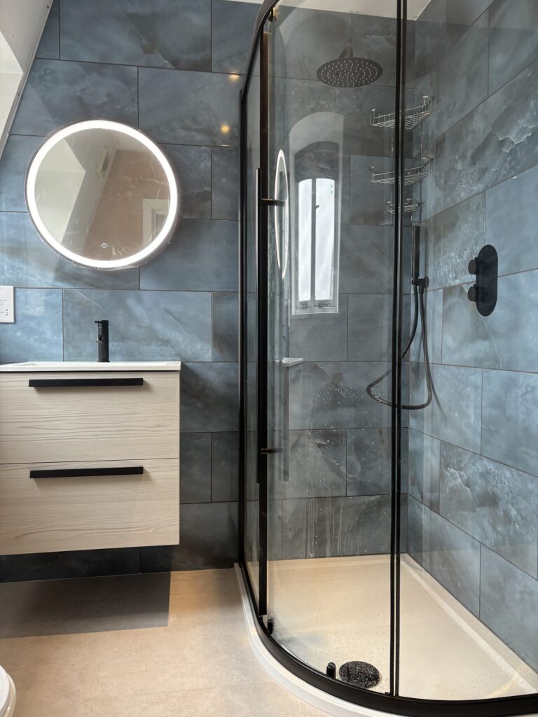 Modern bathroom featuring a black framed shower enclosure, blue porcelain wall tiles, floating vanity unit, round mirror and anthracite heated towel rail installed by JD Designed Bathrooms.