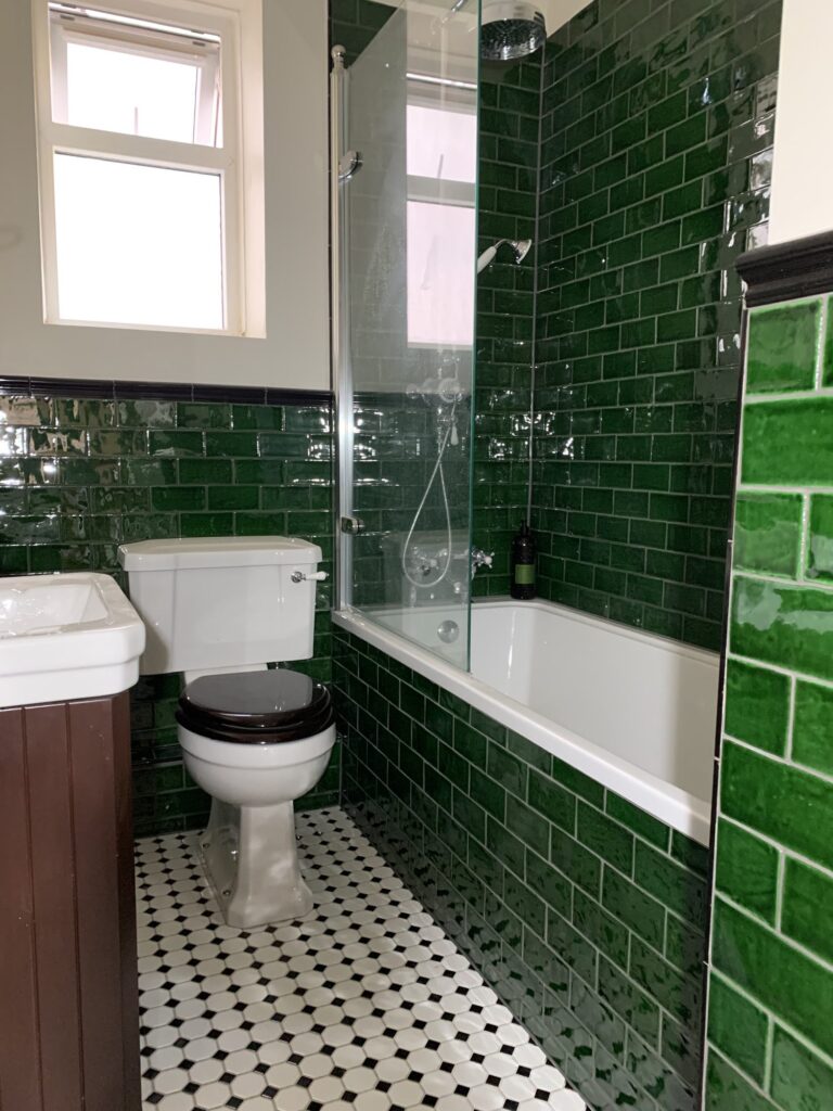 Traditional bathroom featuring glossy green subway tiles, brass shower fittings, white bath with glass screen, Victorian style basin and patterned porcelain floor tiles installed by JD Designed Bathrooms.