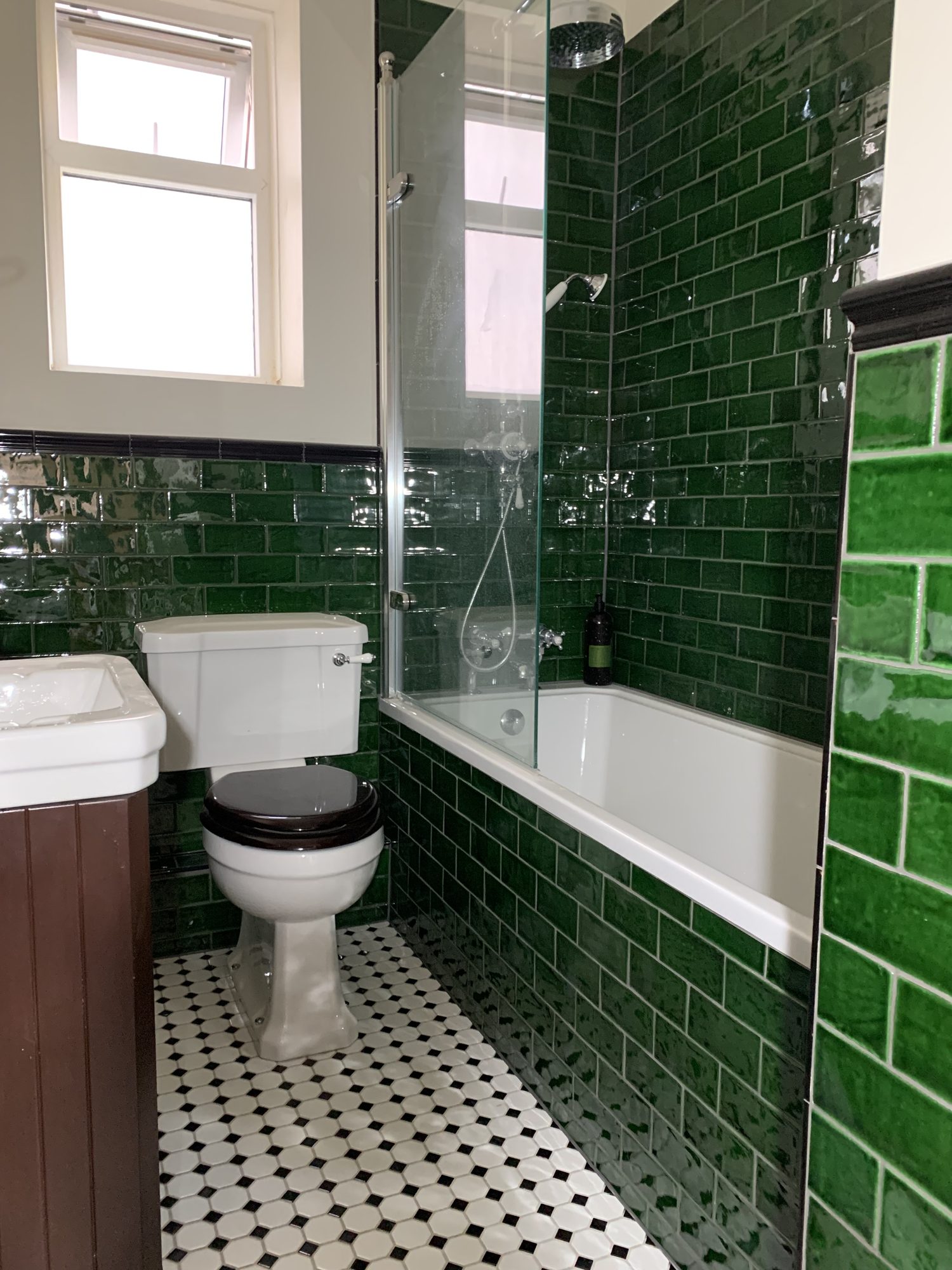Traditional bathroom featuring glossy green subway tiles, brass shower fittings, white bath with glass screen, Victorian style basin and patterned porcelain floor tiles installed by JD Designed Bathrooms.