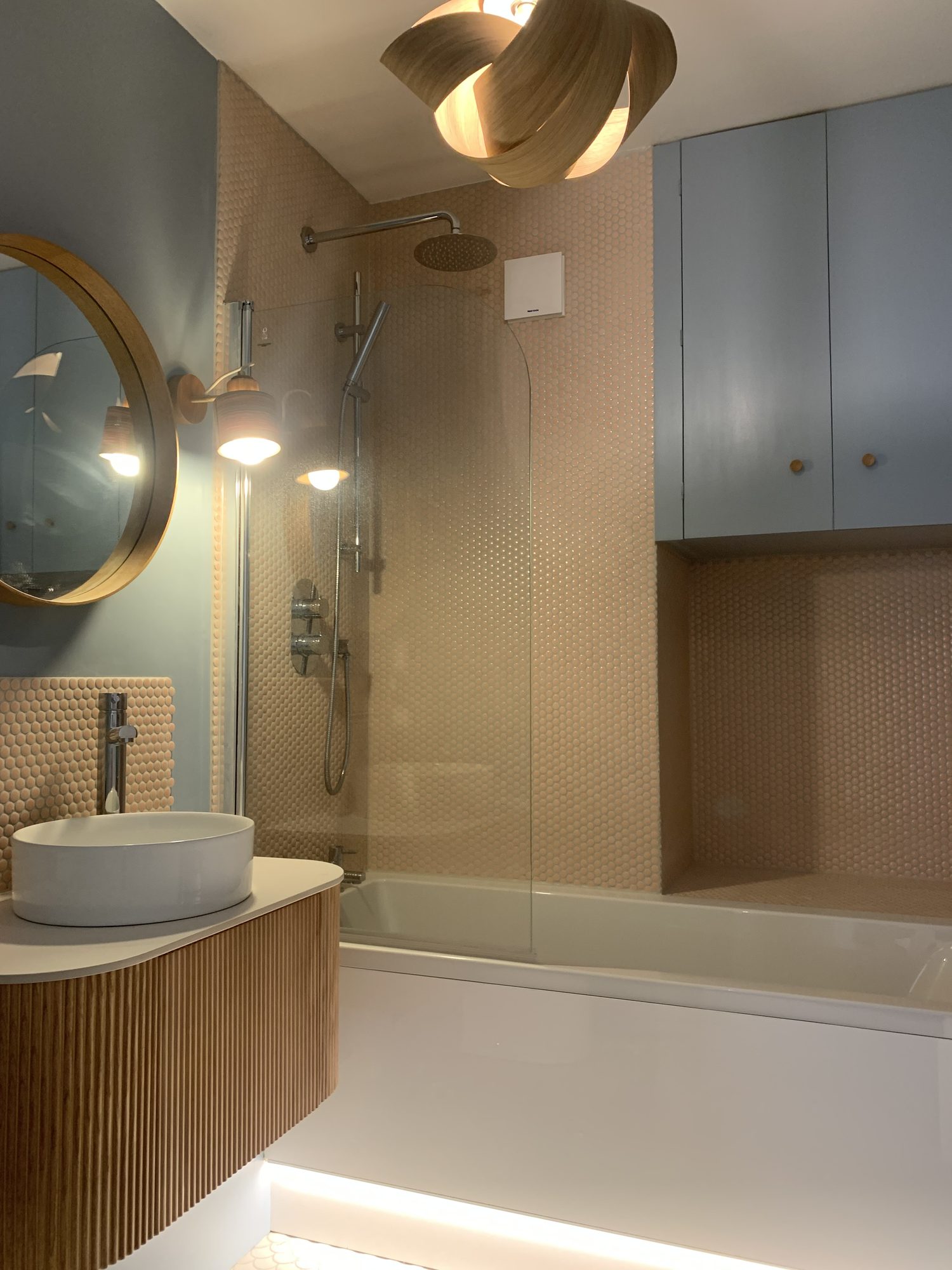 Modern bathroom featuring blush mosaic wall tiles, floating fluted vanity unit, countertop basin, built-in storage, walk-in shower over bath and contemporary feature lighting installed by JD Designed Bathrooms.