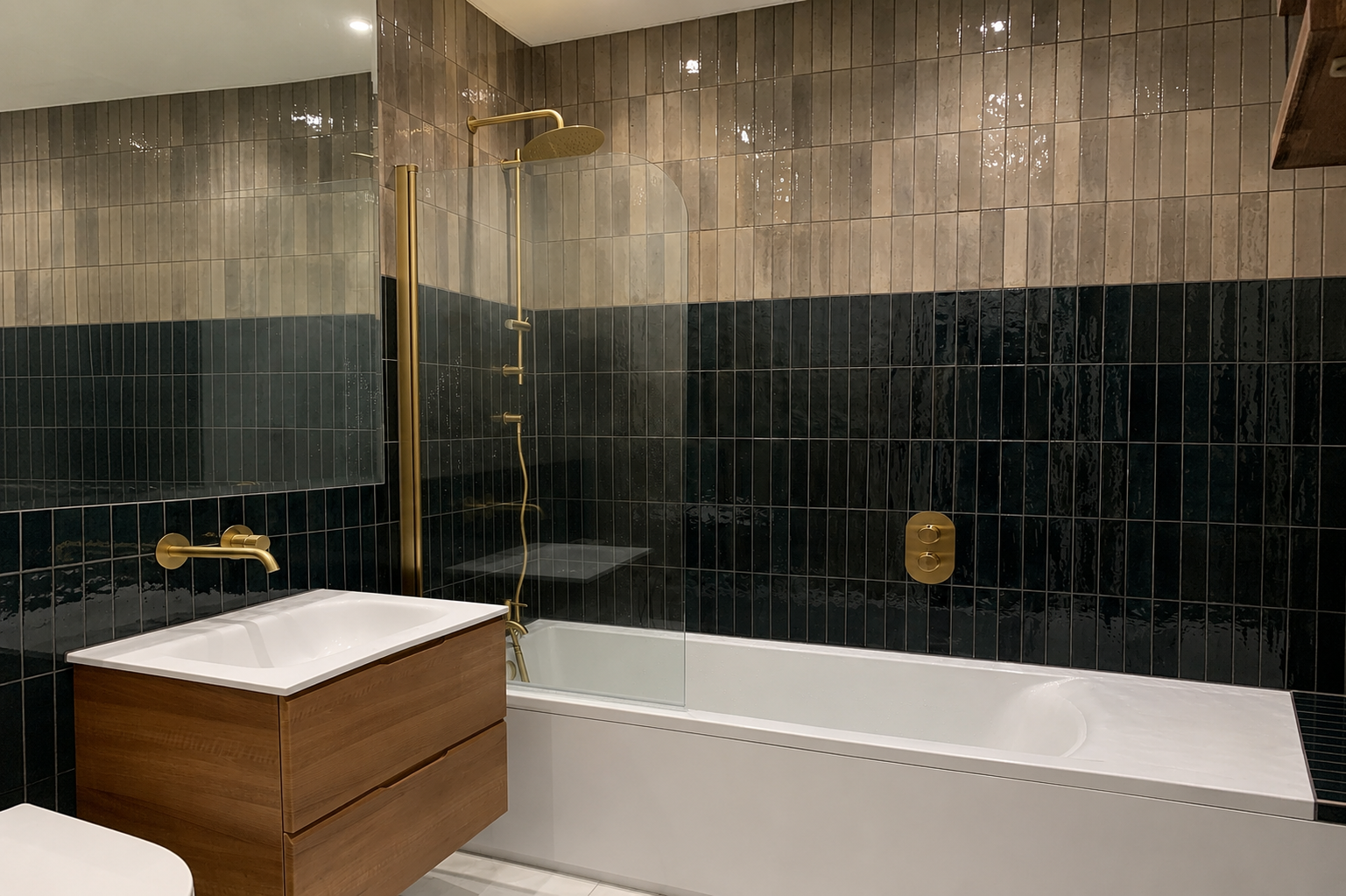 Modern bathroom with two-tone tiled bath, brass rainfall shower, floating wood vanity unit and glass bath screen.